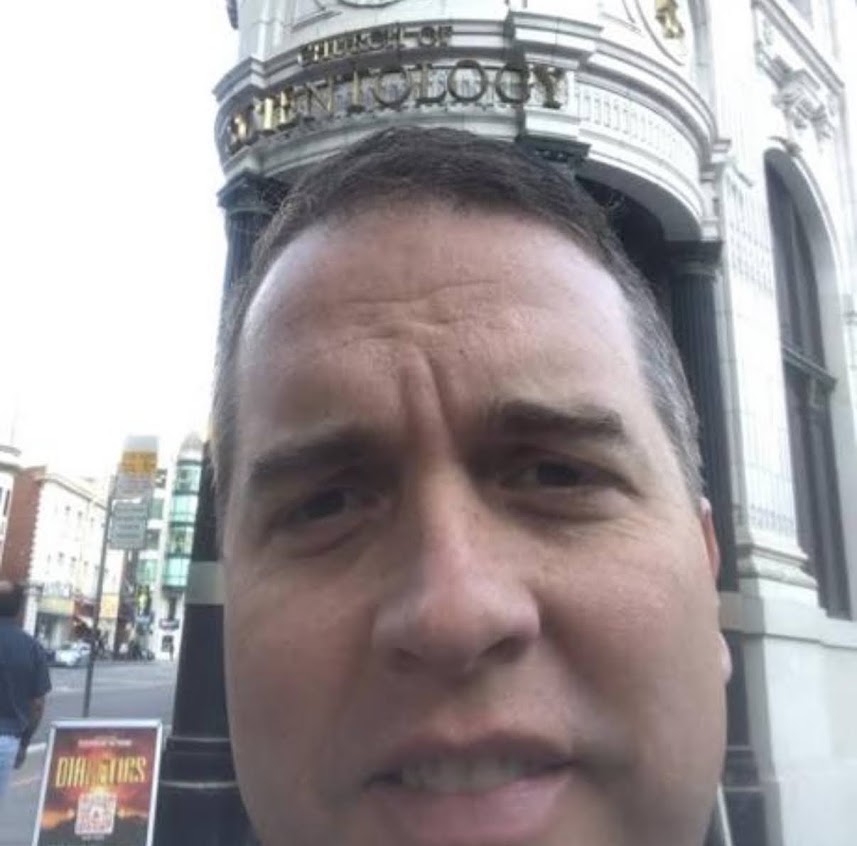 A man taking a determined selfie in front of an ornate Church of Scientology building on a city street corner